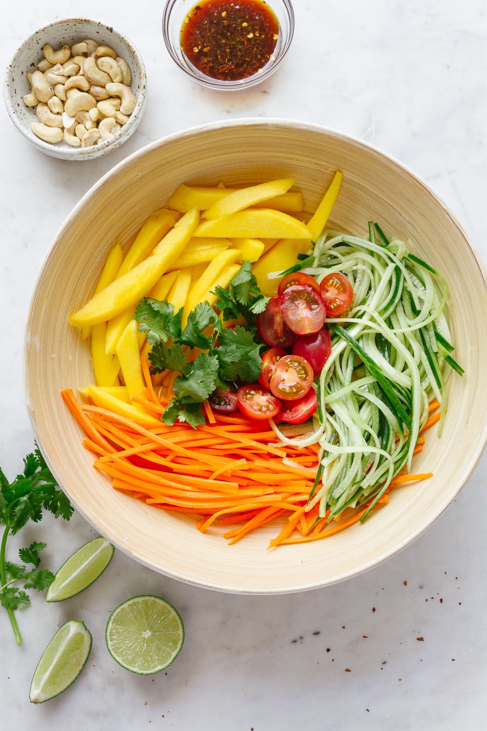 top down view of a bamboo bowl with ingredients of thai mango salad surrounded by cilantro, cashews and dressing in a small glass bowl