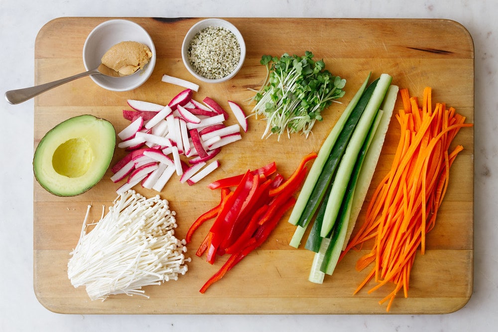 ingredients for vegan sushi roll filling on a wooden cutting board, including miso, avocado, enoki mushrooms, bell pepper, zucchini, carrots, radish and pea shoots