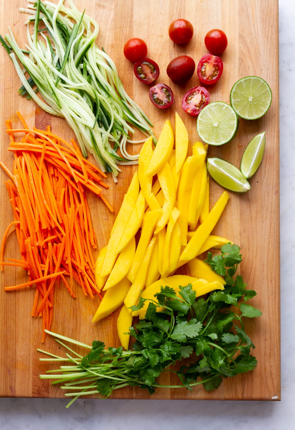 top down view of prepped carrots, cucumber, mango tomatoes, cilantro and lime wedges on a wooden cutting board