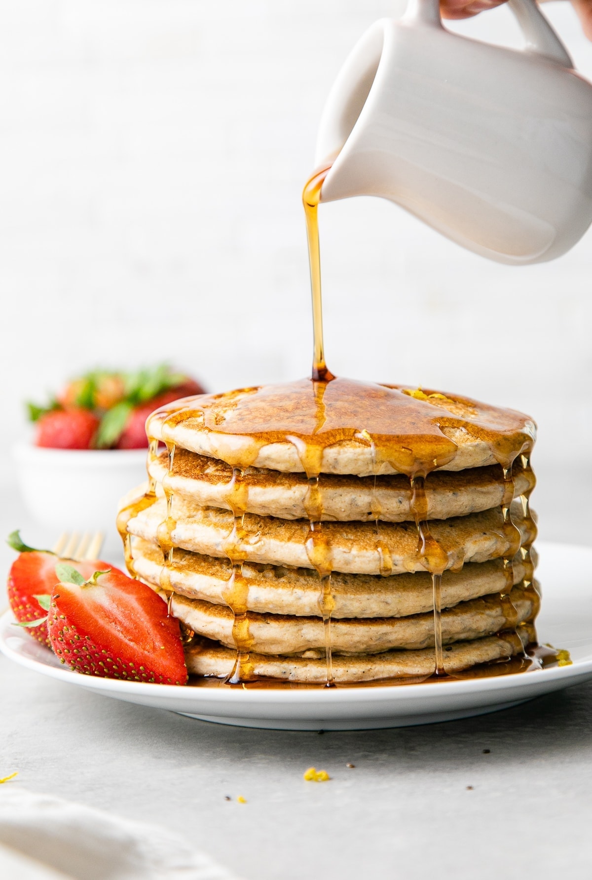 head on view of maple syrup being poured on lemon chia seed pancakes stacked on a small plate.