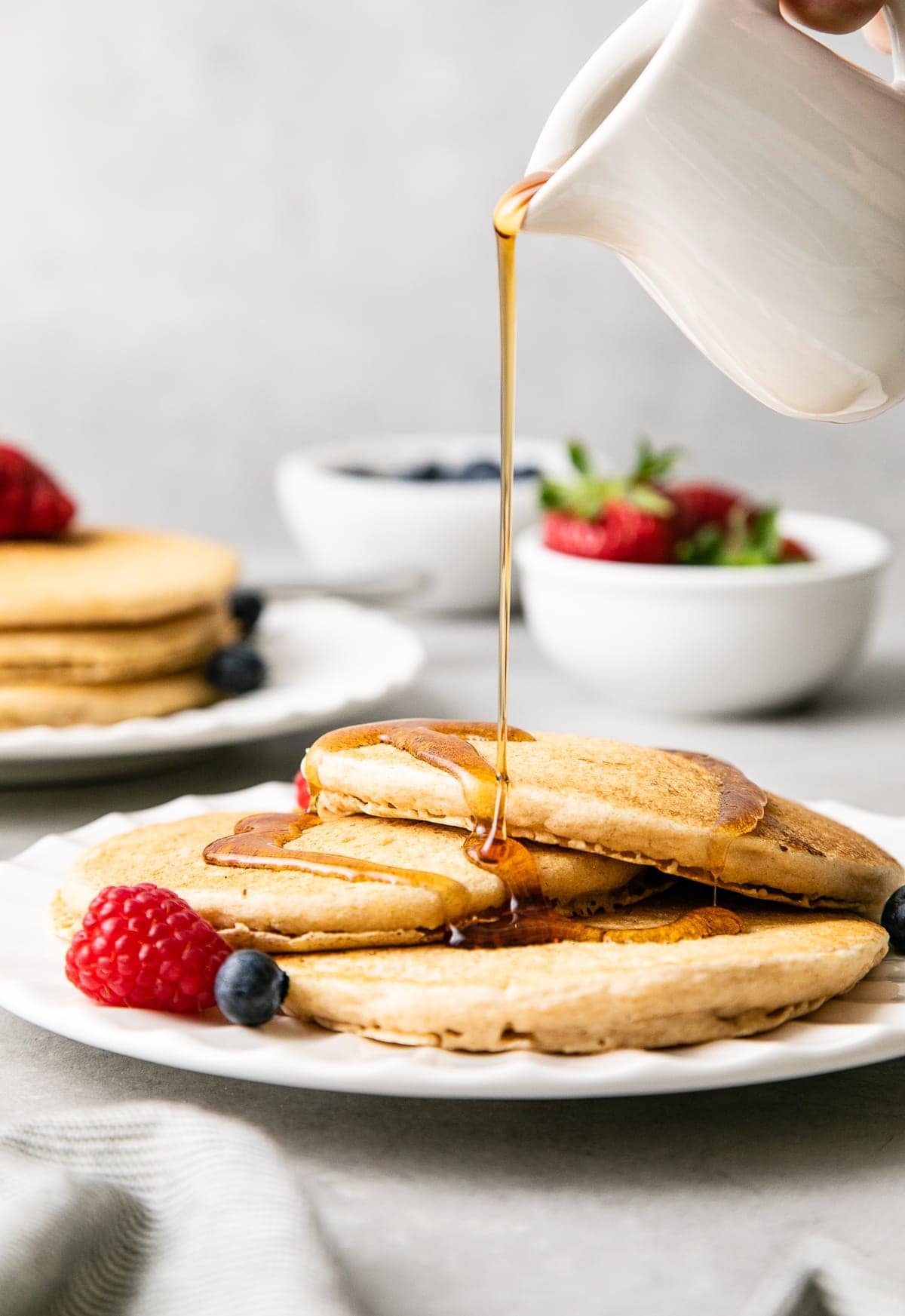 head on view of serving of stacked vegan pancakes on small plate being drizzled with pure maple syrup.