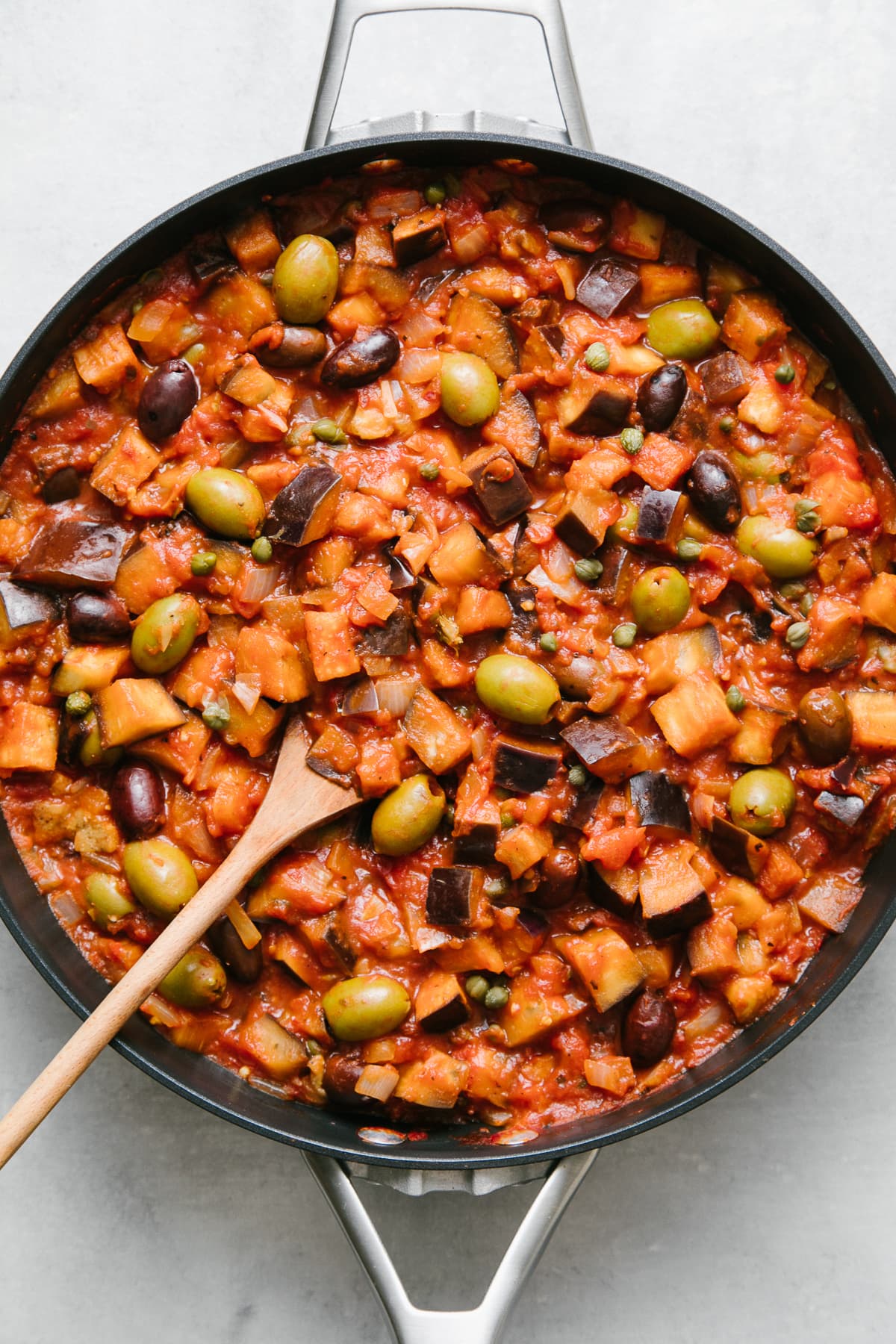 top down view of freshly made eggplant puttanesca in a pan with wooden spoon.