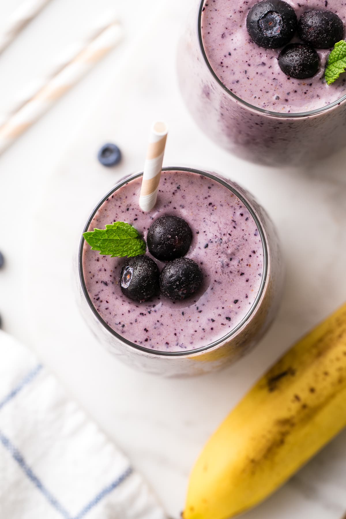 top down view of glass full of blueberry banana smoothie with straw and items surrounding.