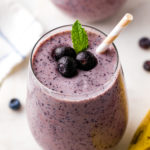side angle view of glass full of blueberry banana smoothie with straw and items surrounding.