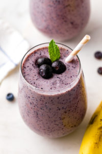 side angle view of glass full of blueberry banana smoothie with straw and items surrounding.
