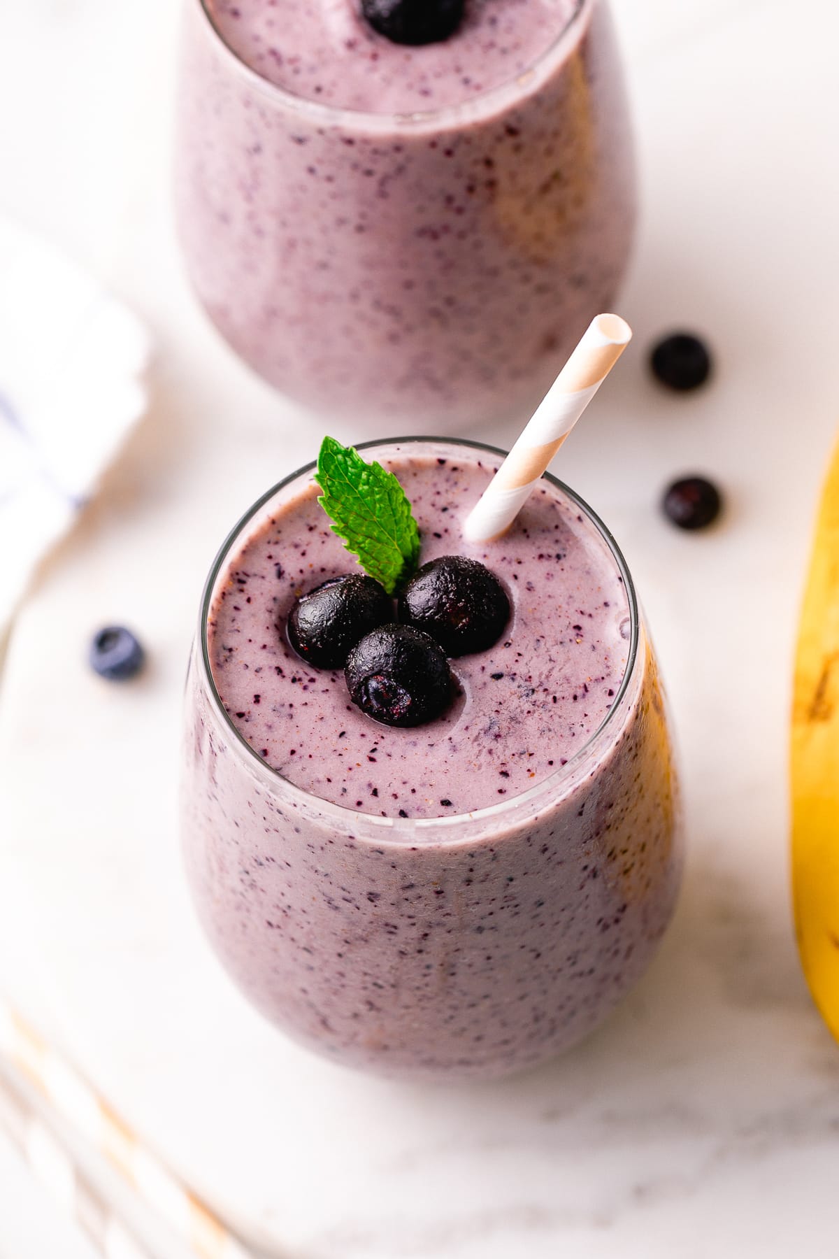 side angle view of glass full of blueberry banana smoothie with straw and items surrounding.