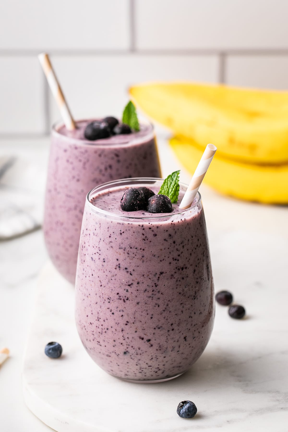 head on view of glass full of blueberry banana smoothie with straw and items surrounding.