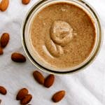 homemade almond butter in a glass jar on white tablecloth with almonds scattered around