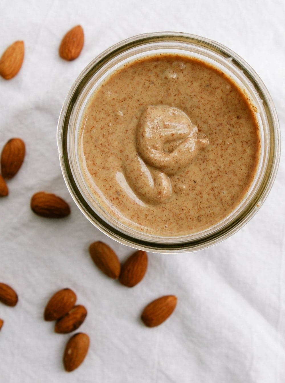 homemade almond butter in a glass jar on white tablecloth with almonds scattered around