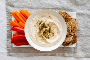 top down view of white bean hummus in a bowl with fresh veggies and chips for dipping on a plate.