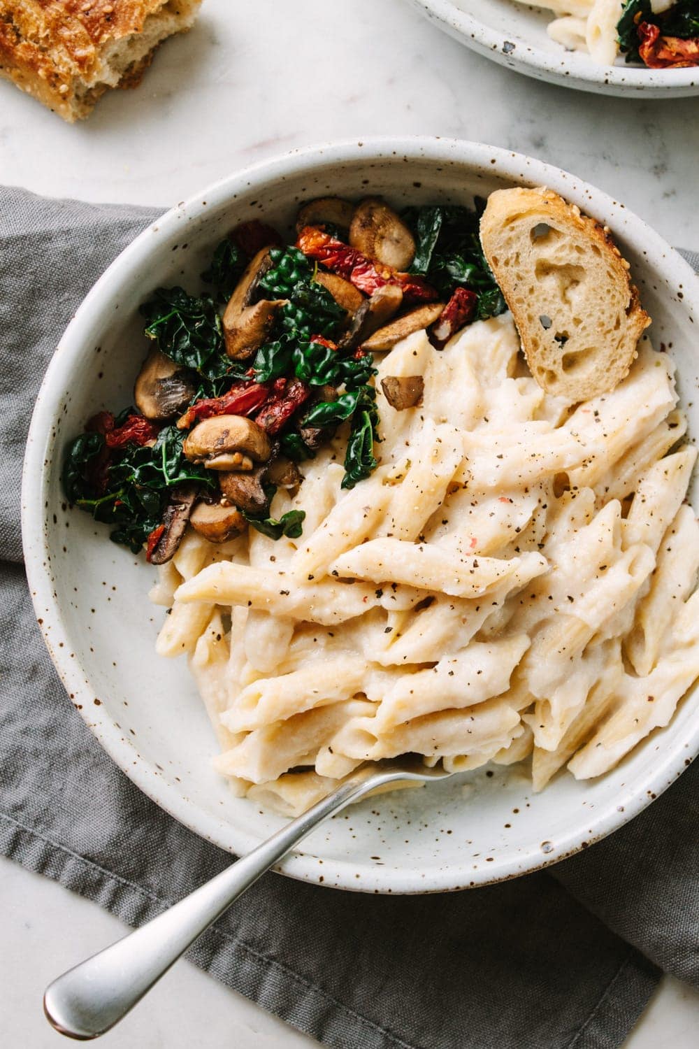 vegan white bean cauliflower alfredo sauce with penne pasta in a bowl with sauteed kale, mushrooms and sun-dried tomatoes.