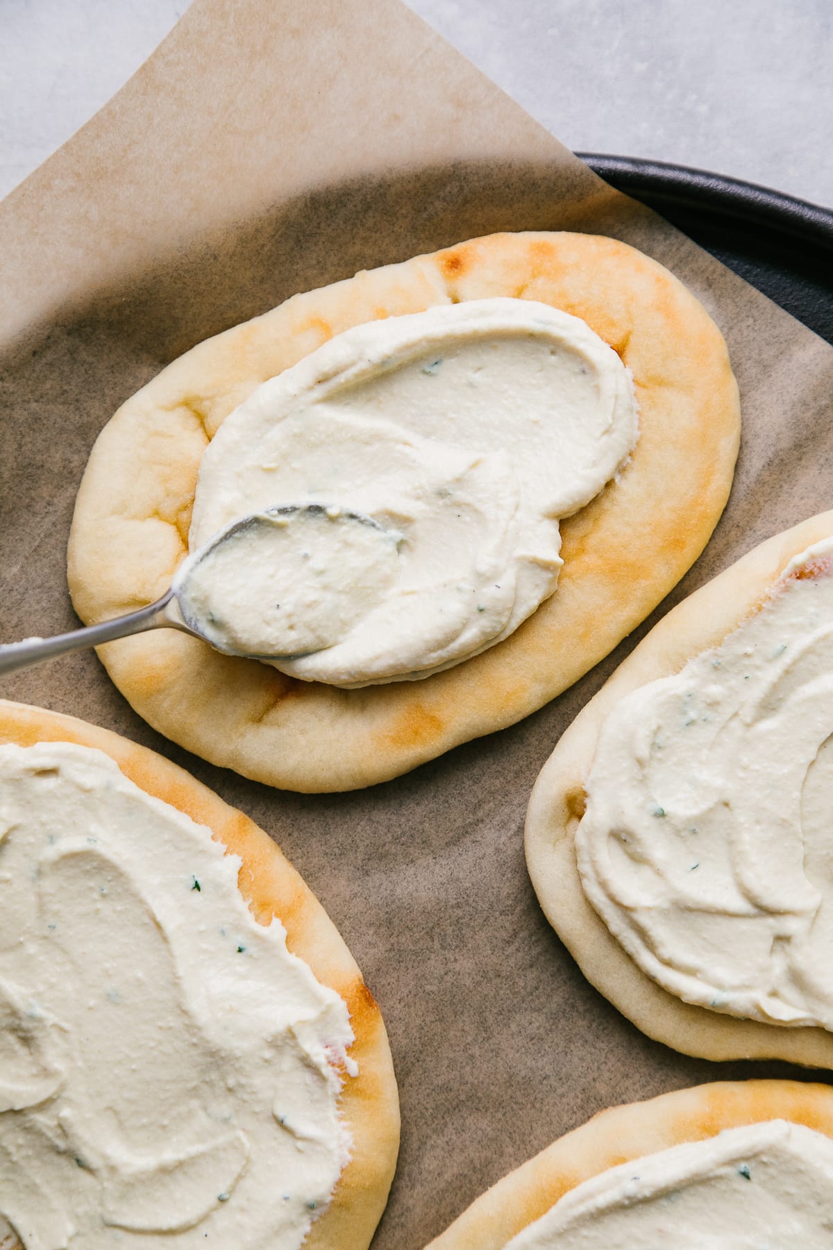 top down view of white pizza sauce being spread on naan flatbread with back of a spoon.