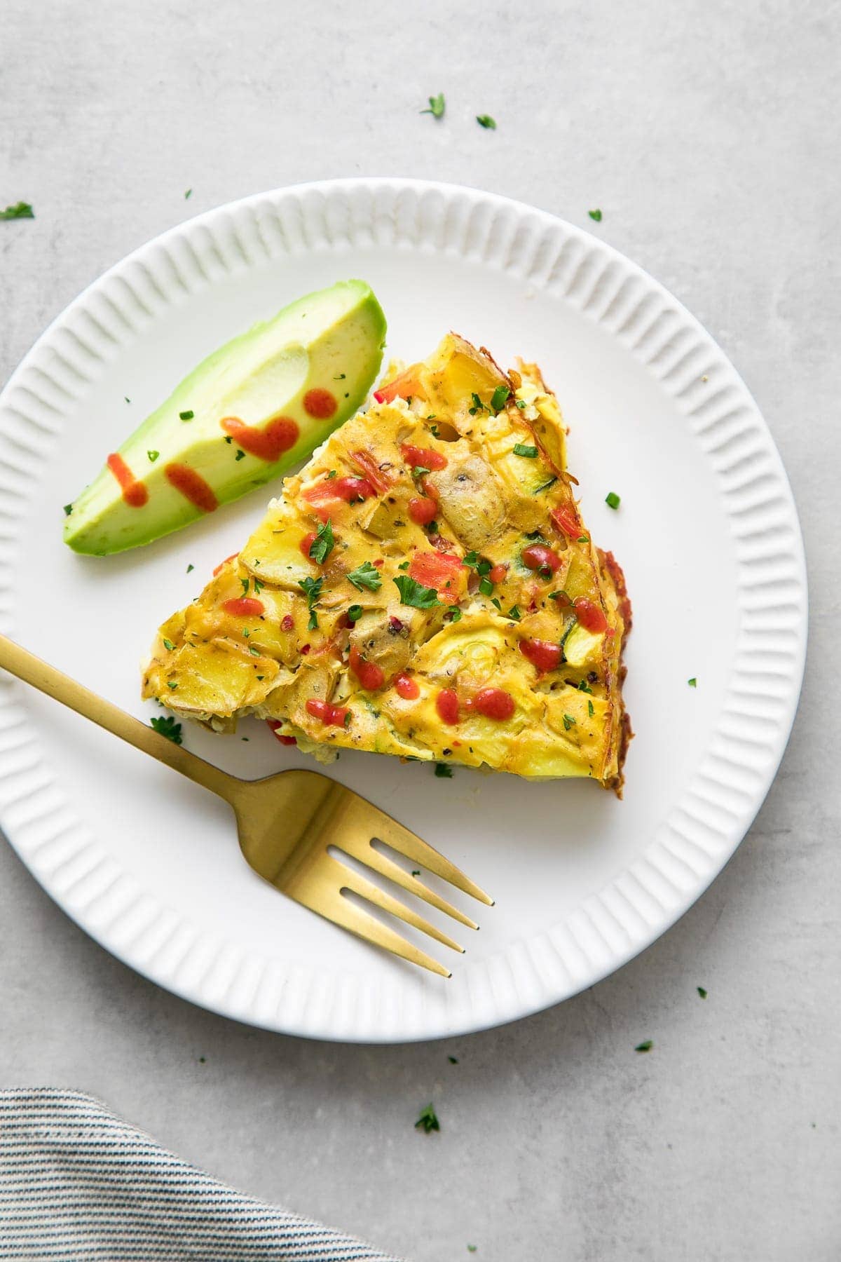 top down view of a slice of vegetable vegan frittata on a small white plate with fork and avocado.
