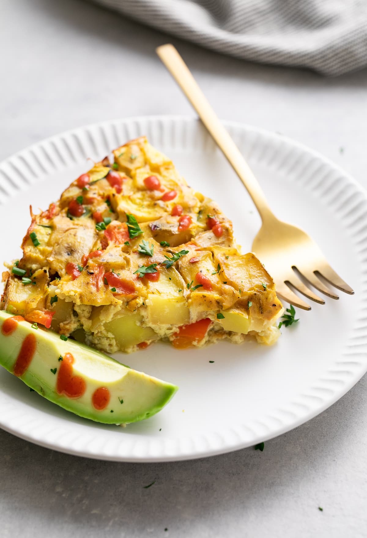 side angle view of a slice of vegetable vegan frittata on a small white plate with fork, avocado and sriracha drizzle.