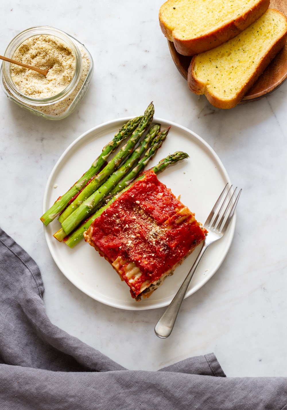top down view of a plate with a slice of simple vegan lasagna and asparagus, with almond parmesan and bread on the side