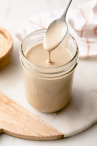 side angle view of tahini dripping from a spoon over jar of tahini with items surrounding.