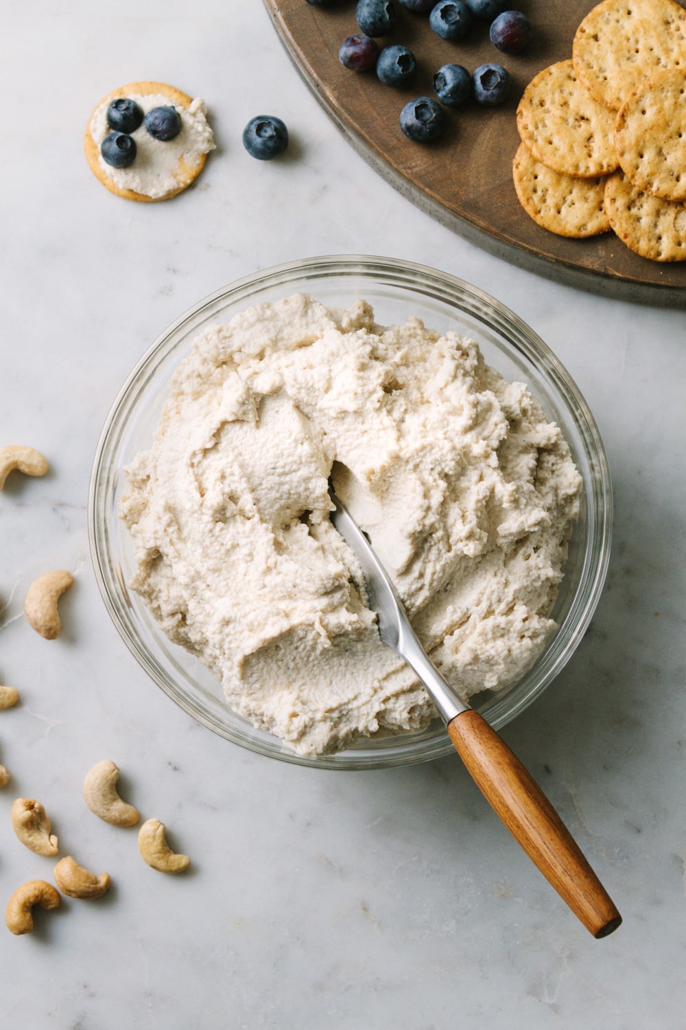 top down view of vegan cashew ricotta cheese in a bowl with a knife with wooden handle, and crackers with blueberries scattered around