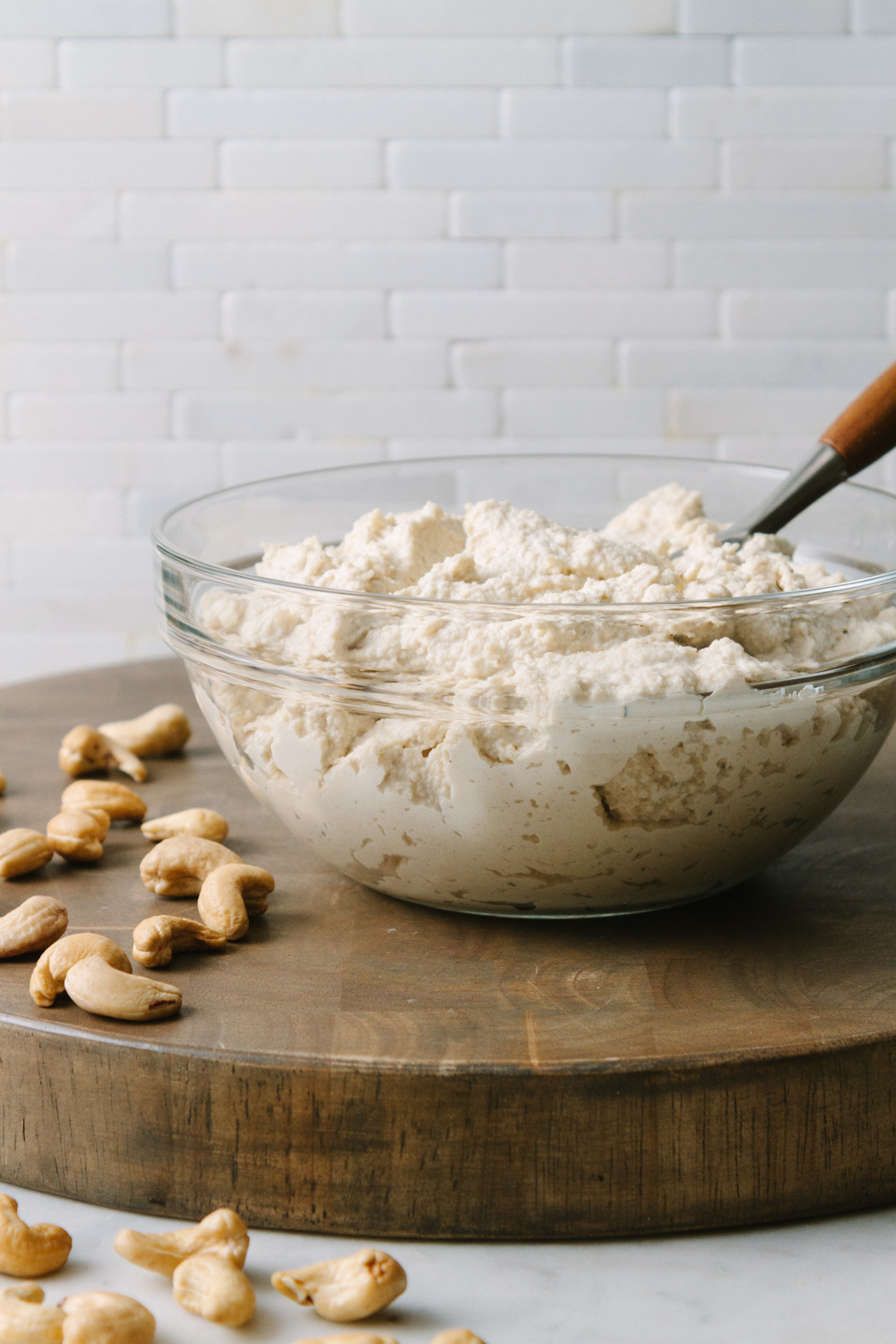 head on view of vegan cashew ricotta cheese in a bowl sitting on a brown wooden cutting board with cashews scattered around