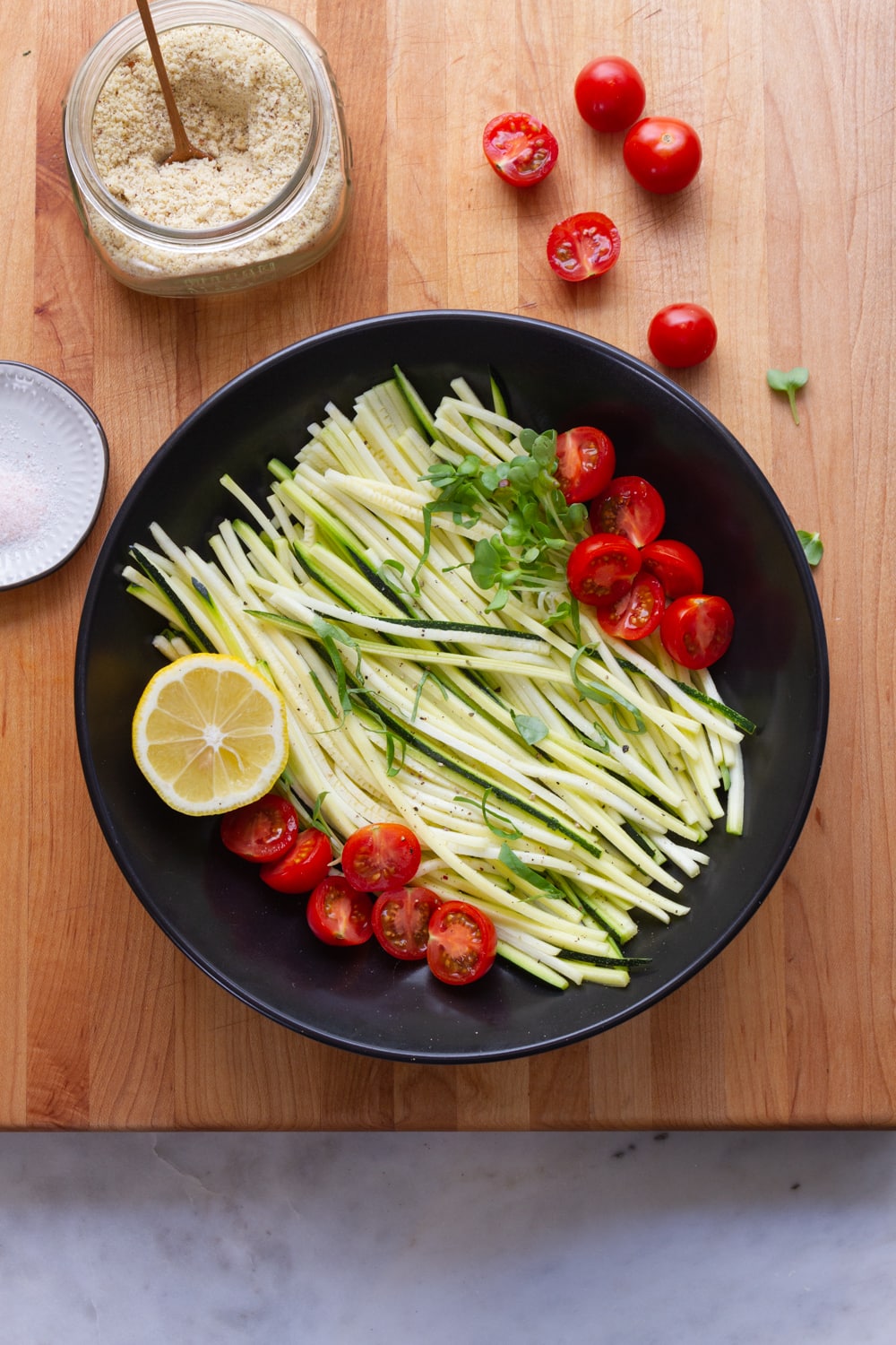 top down view of black bowl filled zucchini pasta, tomatoes, lemon and micro sprouts on a wooden cutting board