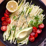 zucchini pasta with tomatoes, avocado, lemon, micro sprouts and almond parmesan in a black bowl on a wooden cutting board