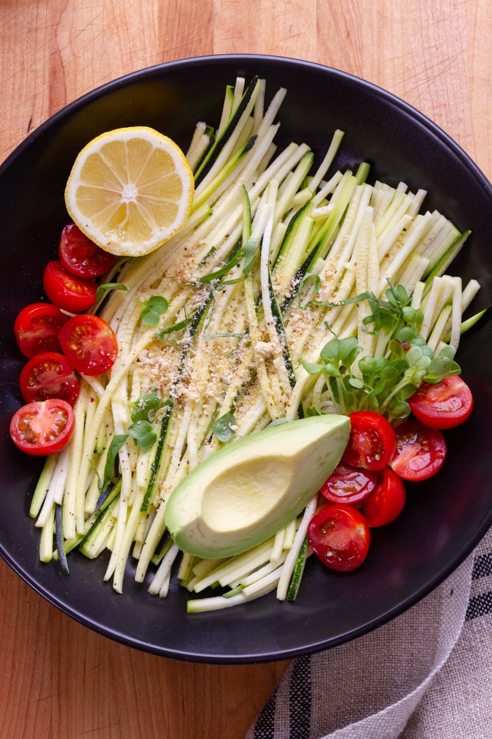zucchini pasta with tomatoes, avocado, lemon, micro sprouts and almond parmesan in a black bowl on a wooden cutting board