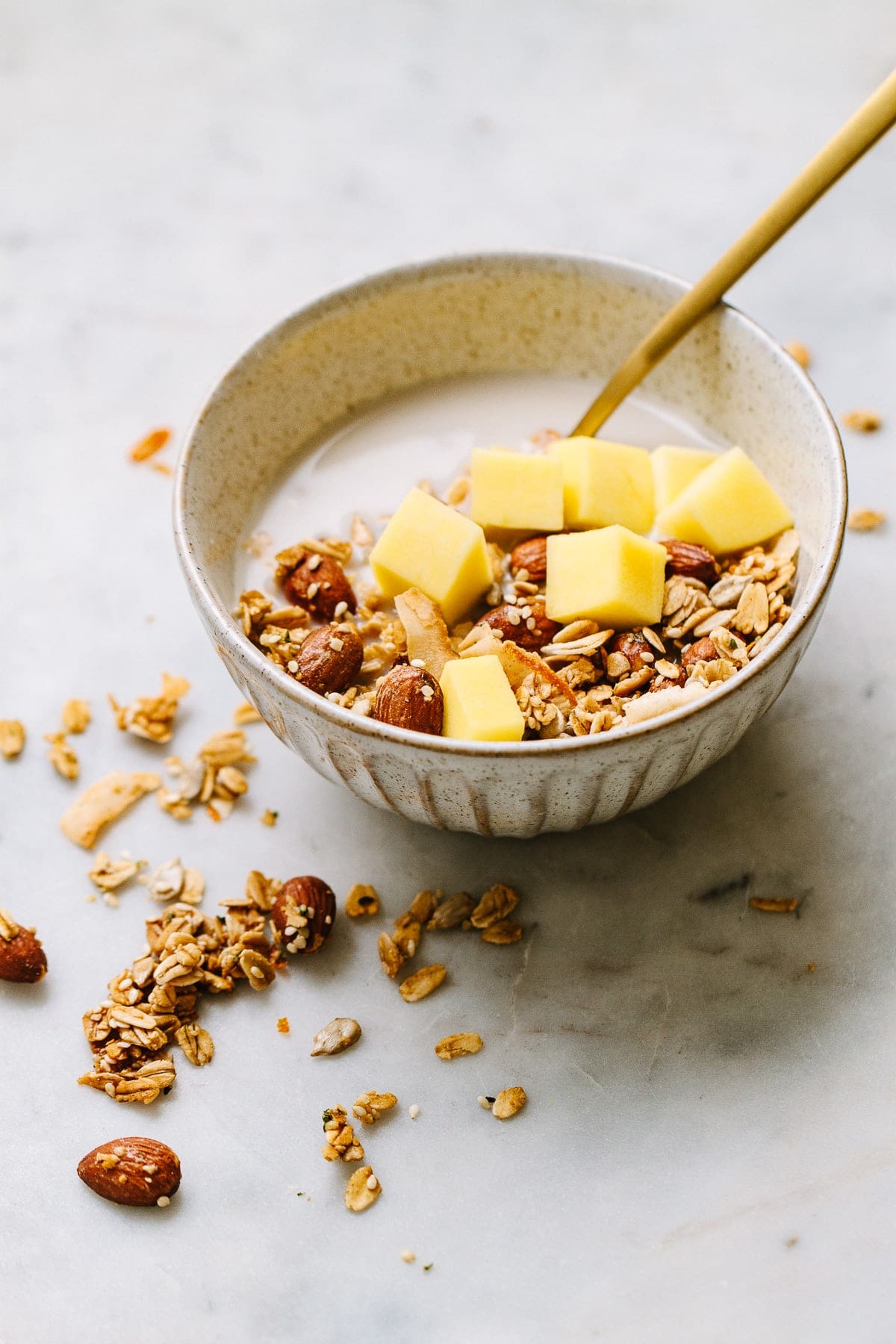 side angle view of a small bowl with a serving of healthy orange ginger granola with cashew milk and diced mango with gold spoon.