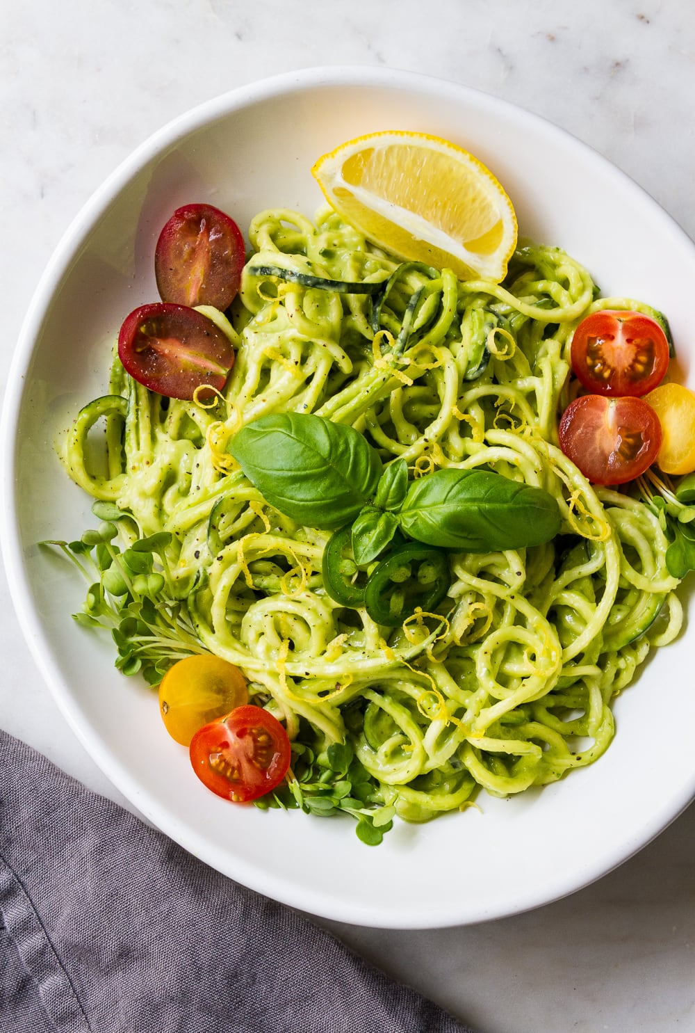 top down view of zucchini pasta with creamy avocado cucumber sauce, sliced cherry tomatoes, micro sprouts, basil, lemon zest and sliced jalapeno in a white bowl with grey napkin