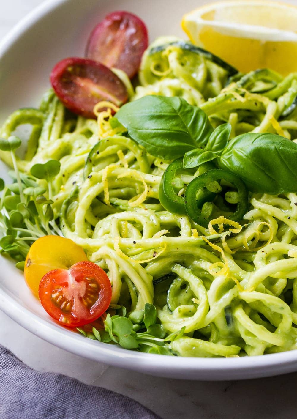 close up, side angle view of zucchini pasta with creamy avocado cucumber sauce, sliced cherry tomatoes, micro sprouts, basil, lemon zest and sliced jalapeno in a white bowl with grey napkin