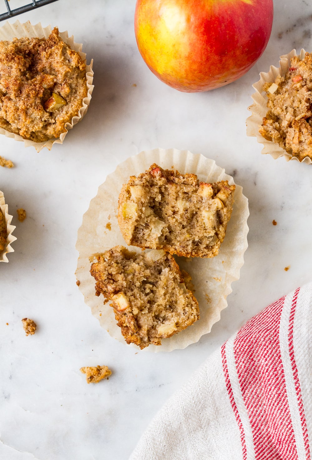 top down view of an apple cinnamon muffin pulled apart, laying on it paper liner, surrounded by more muffins, an apple, and cream and red striped dishtowel 