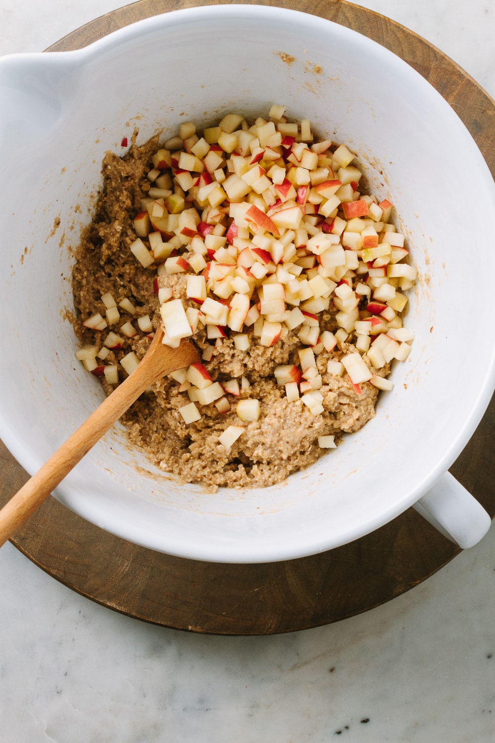 top down view of white mixing bowl with apple cinnamon muffin batter mixed and finely diced apples added before mixing, on a round wooden cutting board