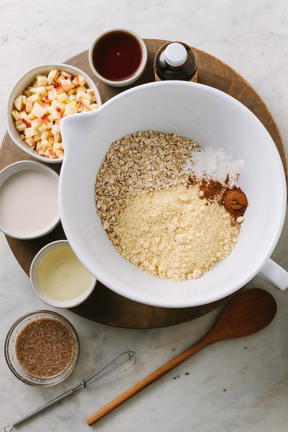 top down view of a mixing bowl filled with almond flour, oats, baking powder, salt and cinnamon, surrounded by small bowls filled with finely diced apples, plant milk, pure maple syrup, oil, flax egg, and bottle of vanilla on a circular brown wooden cutting board