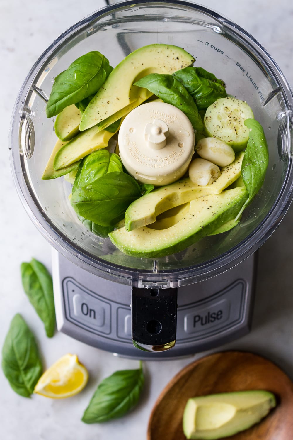 top down view of food processor with sliced avocado, cucumbers, basil leaves, salt, garlic, pepper and lemon juice before putting the lid on to process into a creamy sauce