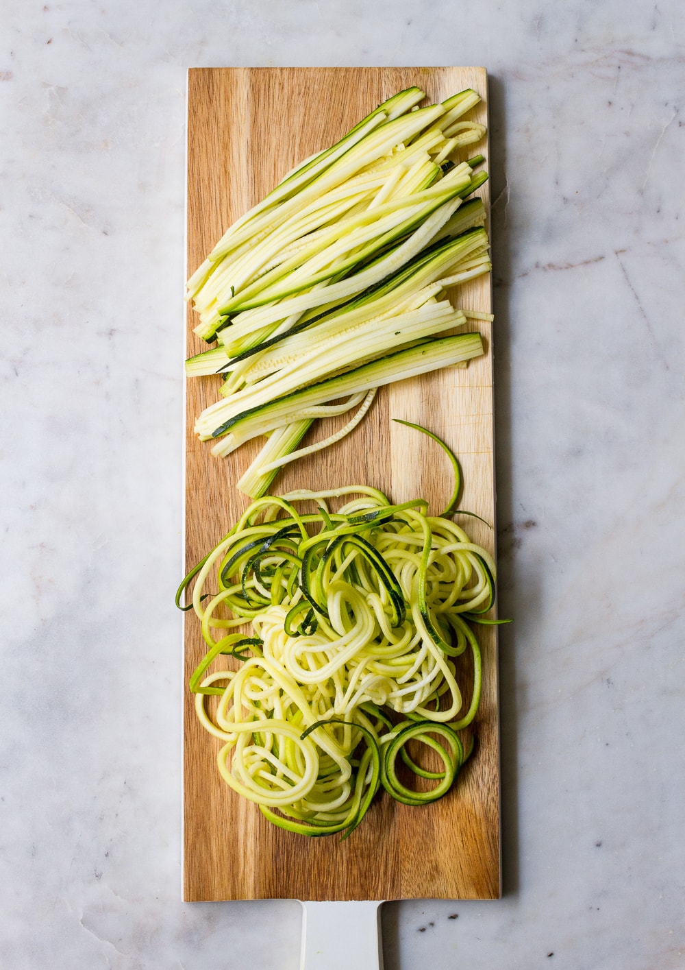 top down view of julienned zucchin and spiralized zuccini on a long rectangular wooden serving paddle board with white handle, on a marble slab