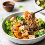 side angle view of chopsticks holding a bite of sesame soba noodle salad with tofu with items surrounding.