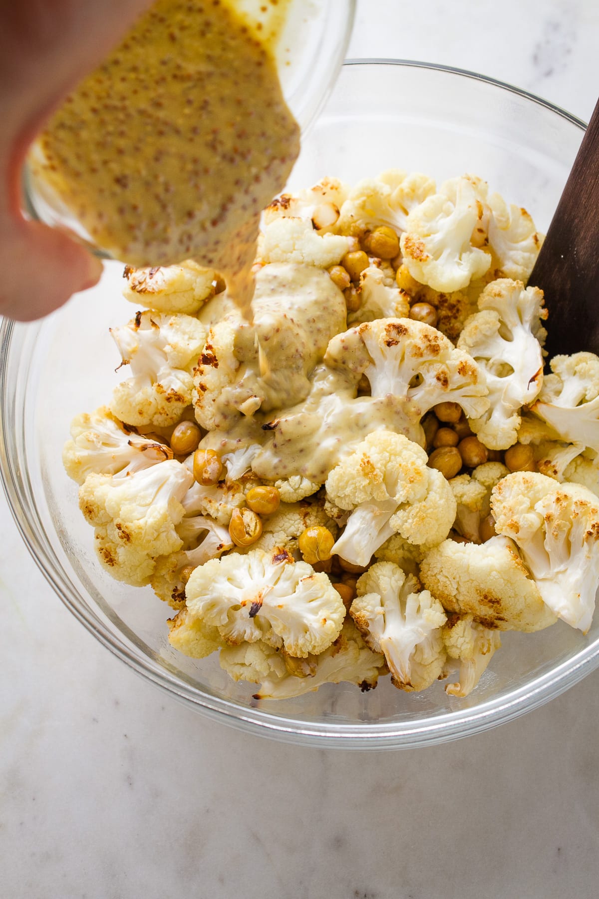 top down view of roasted cauliflower and chickpeas in a glass mixing bowl with mustard dressing poured overtop.