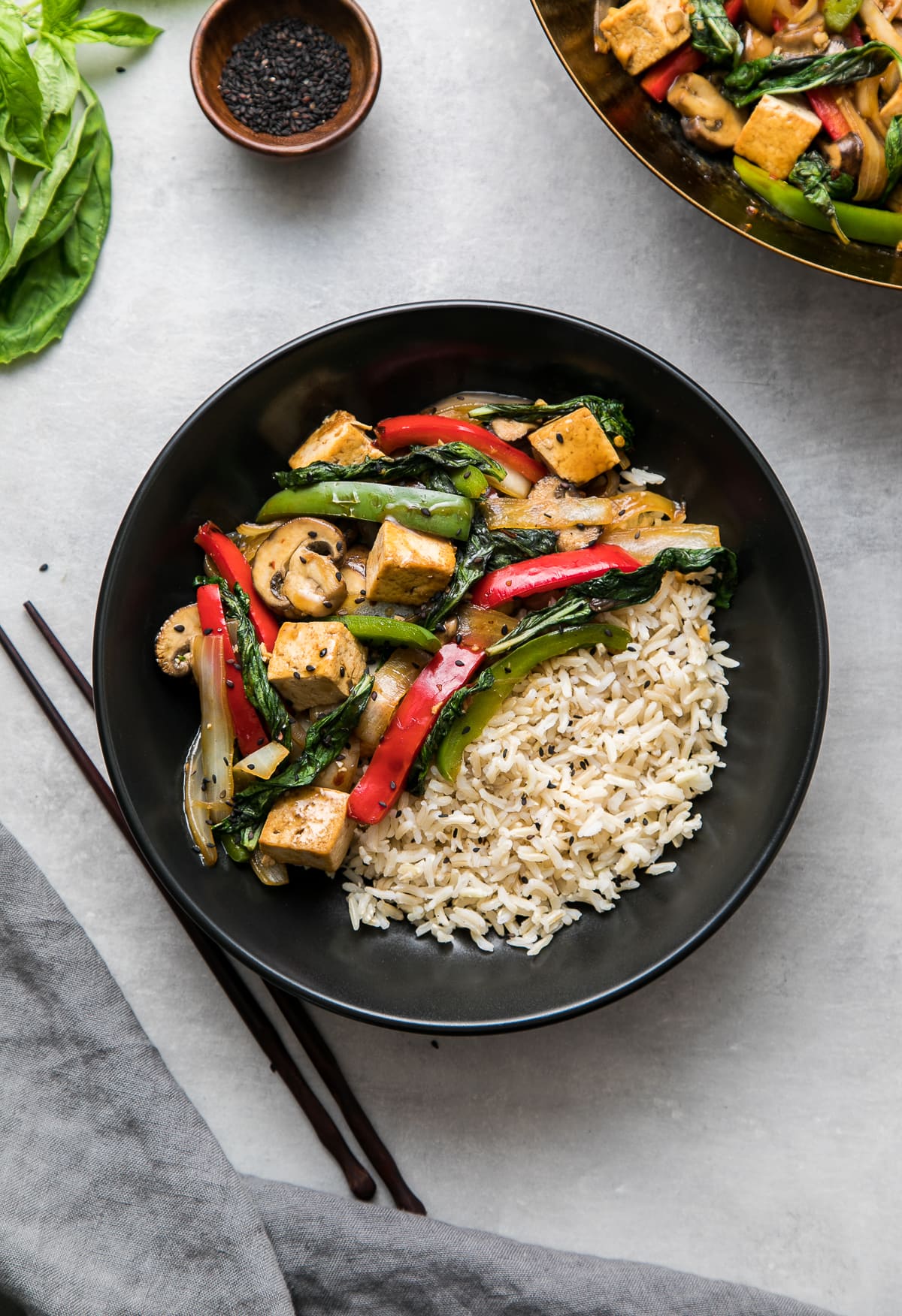 top down view of a serving of thai basil stir fry with tofu and veggies, and rice on the side, in a black bowl.