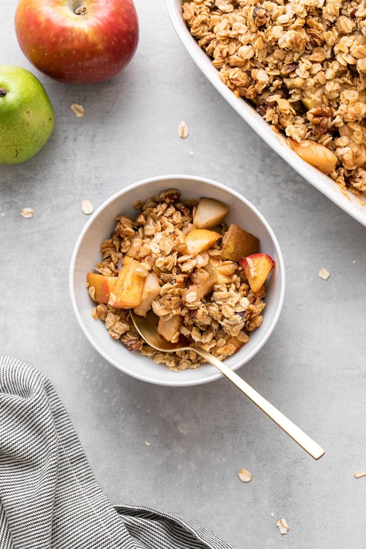 top down view of a serving of healthy apple pear crisp in a white serving bowl with spoon and items surrounding.