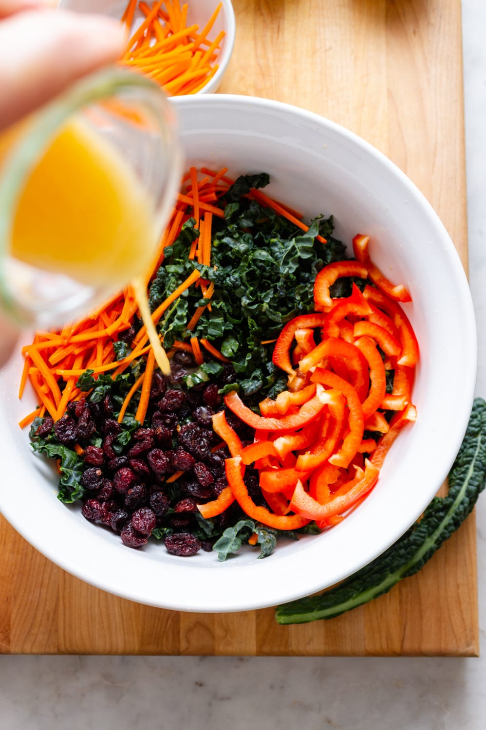 top down view of orange dressing being poured into a white bowl filled with chopped kale, shredded carrots, dried cranberries and sliced red bell peppers.