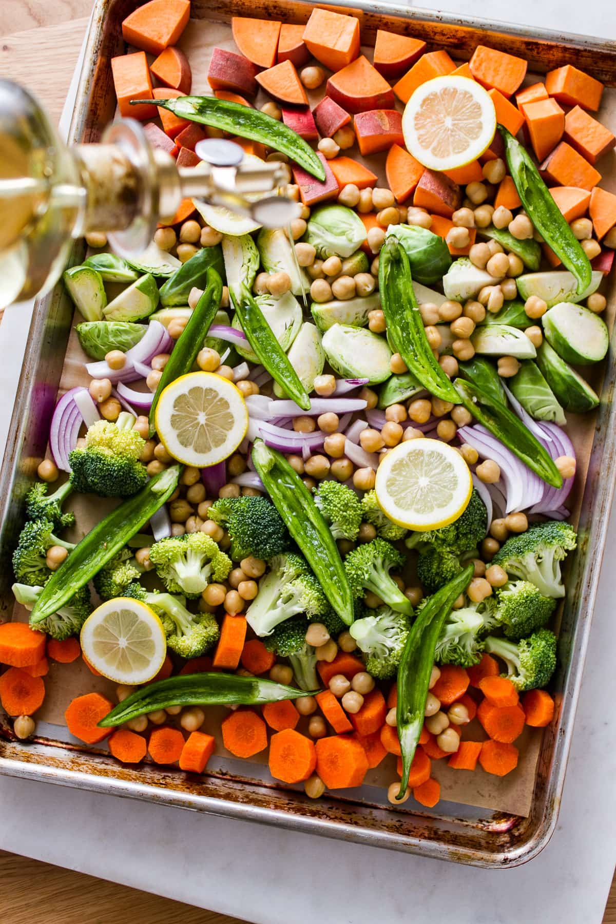 top down view of prepped vegetables on a rimmed baking sheet being drizzled with olive oil.