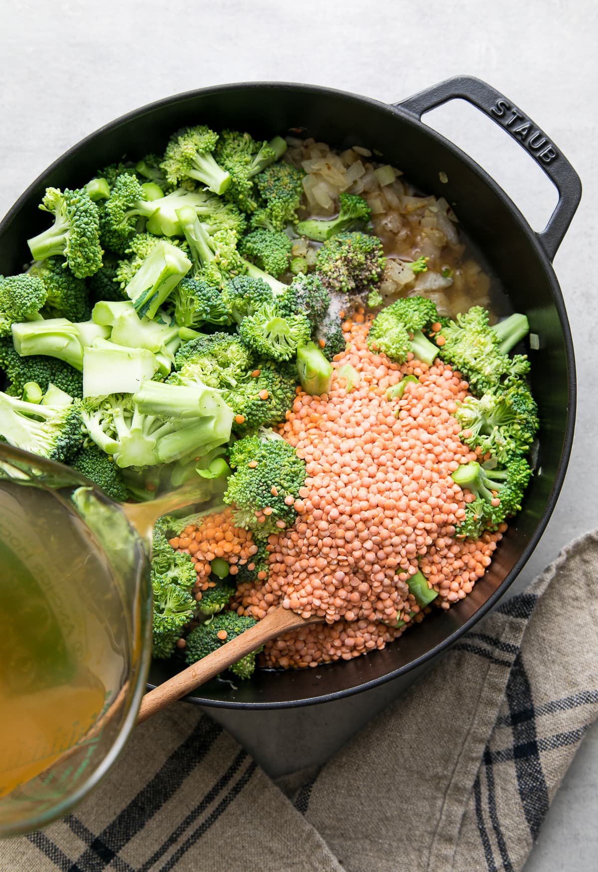 top down view showing the process of adding vegetable broth to pot filled with broccoli red lentil ingredients.