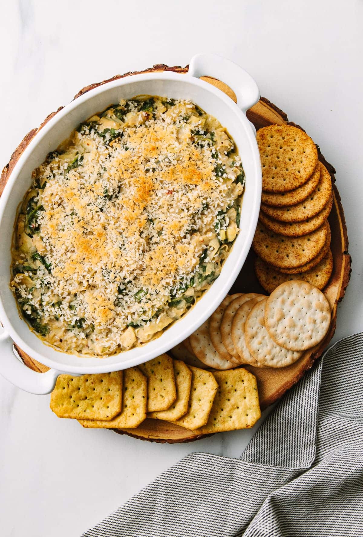 top down view showing the process of adding breadcrumbs to spinach artichoke white bean dip after baking with crackers surrounding.