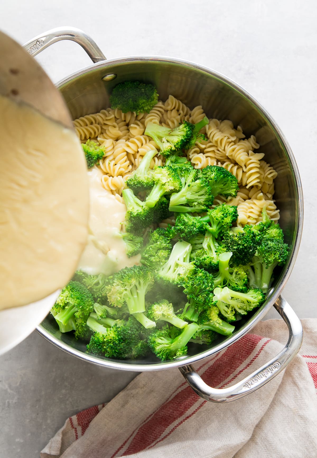 top down view showing the process of pouring creamy vegan sauce into pot of cooked pasta and broccoli.