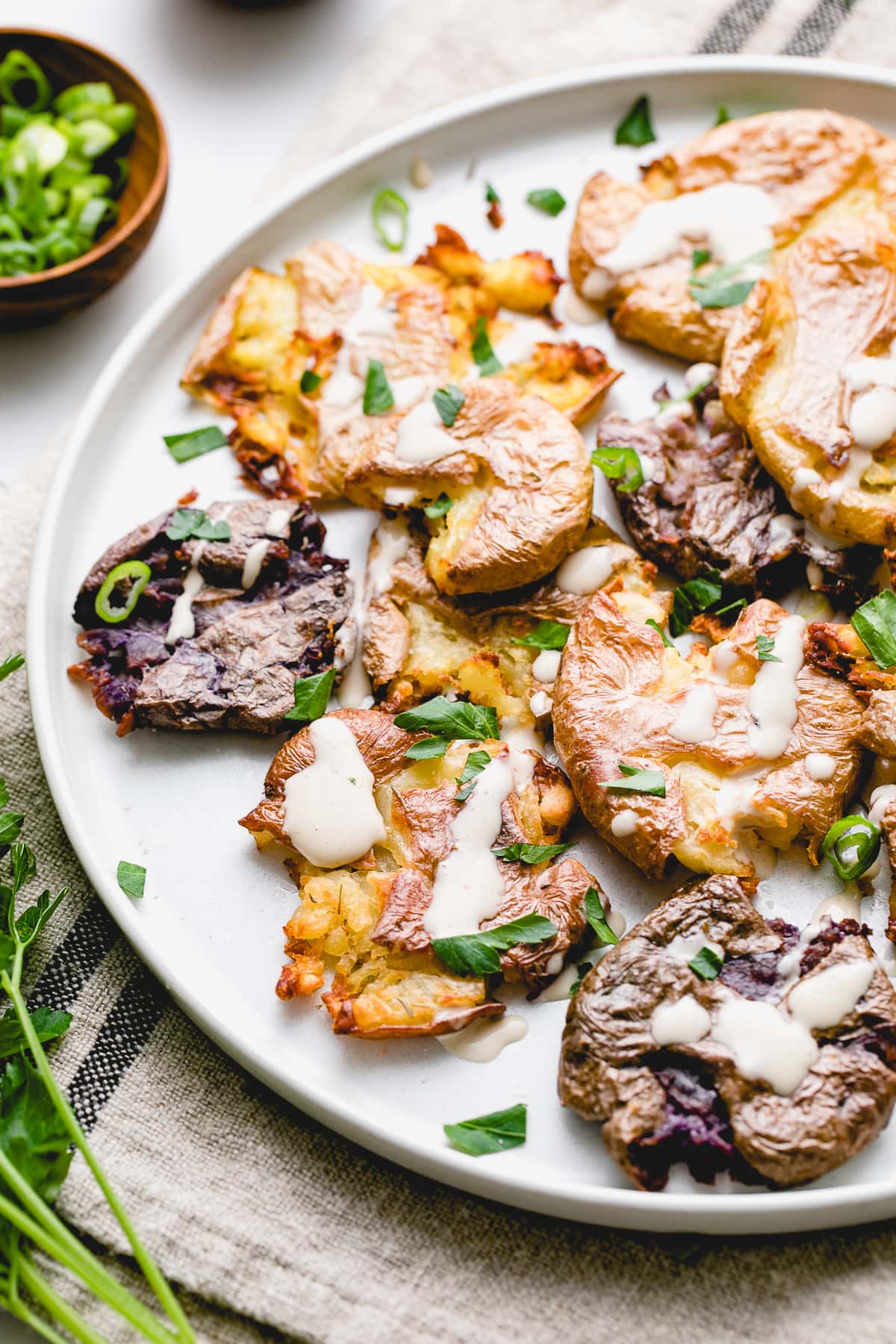 side angle view of plated crispy smashed potatoes with tahini drizzle and items surrounding.