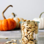 head on view of roasted pumpkin seeds in a glass jar with items surrounding.