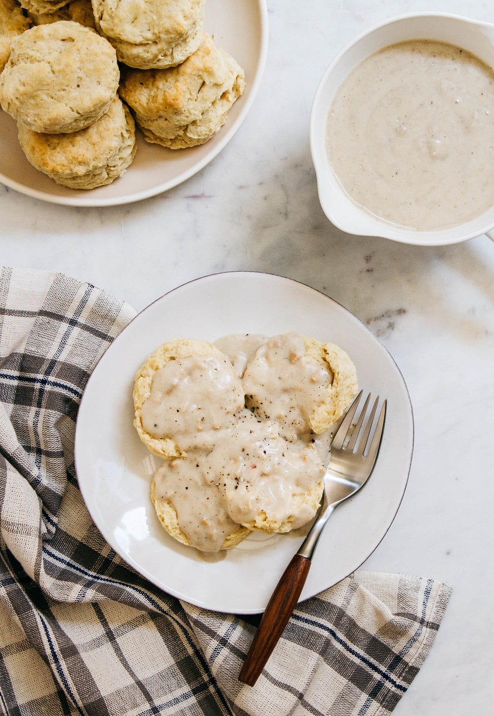 top down view of a plate of biscuits and gravy with fork, surrounded by a plate of stacked biscuits and gravy bowl.