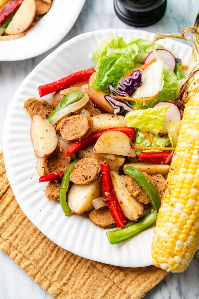 top down view of plated vegan kielbasa sausage with peppers and potatoes with side of corn and salad on a white plate with items surrounding.