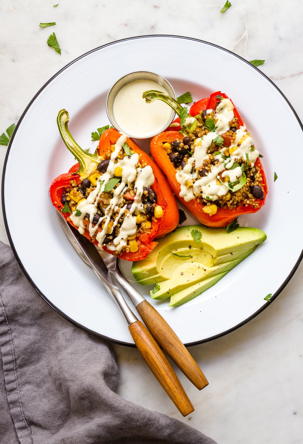 top down view of 2 halves of roasted bell pepper stuffed with tex-mex quinoa and avocado on the side on a white plate with black rim and fork and knife.