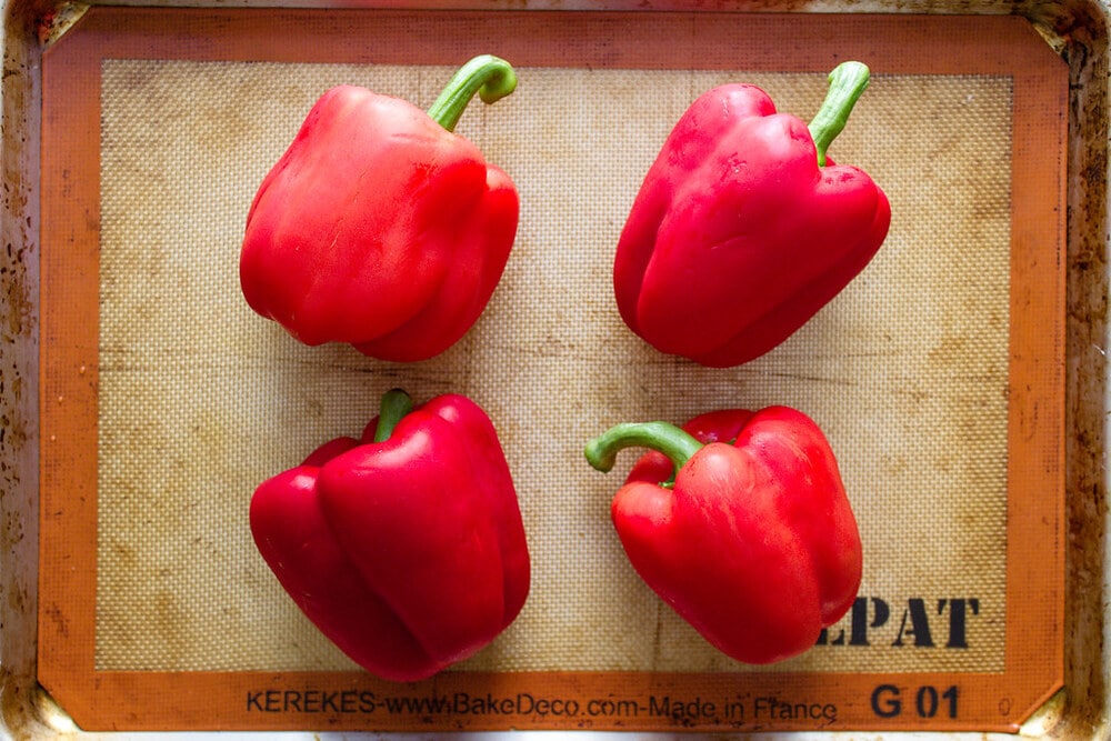 top down view of whole red peppers on a rimmed baking sheet ready to be roasted in the oven.