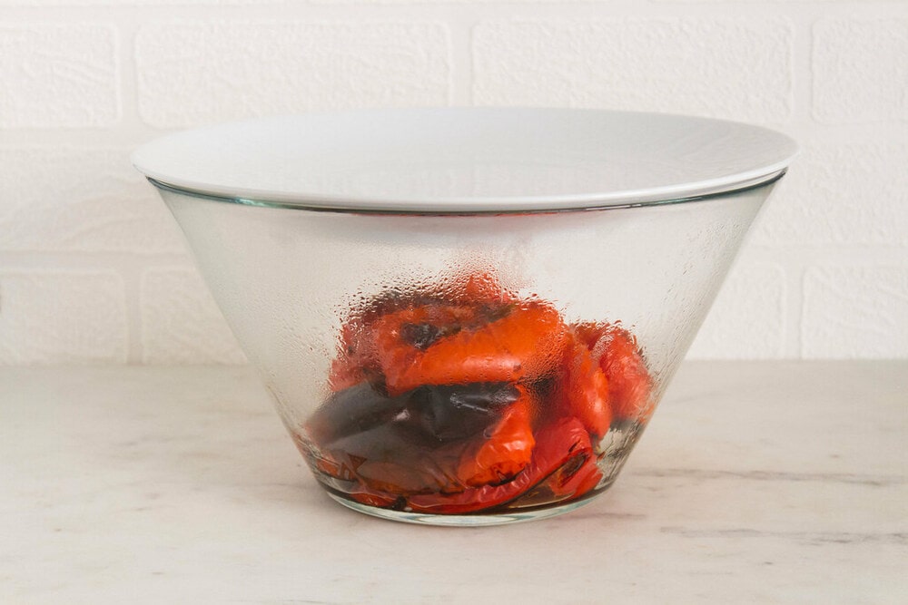 roasted red peppers steaming in a glass bowl covered with a small white plate.