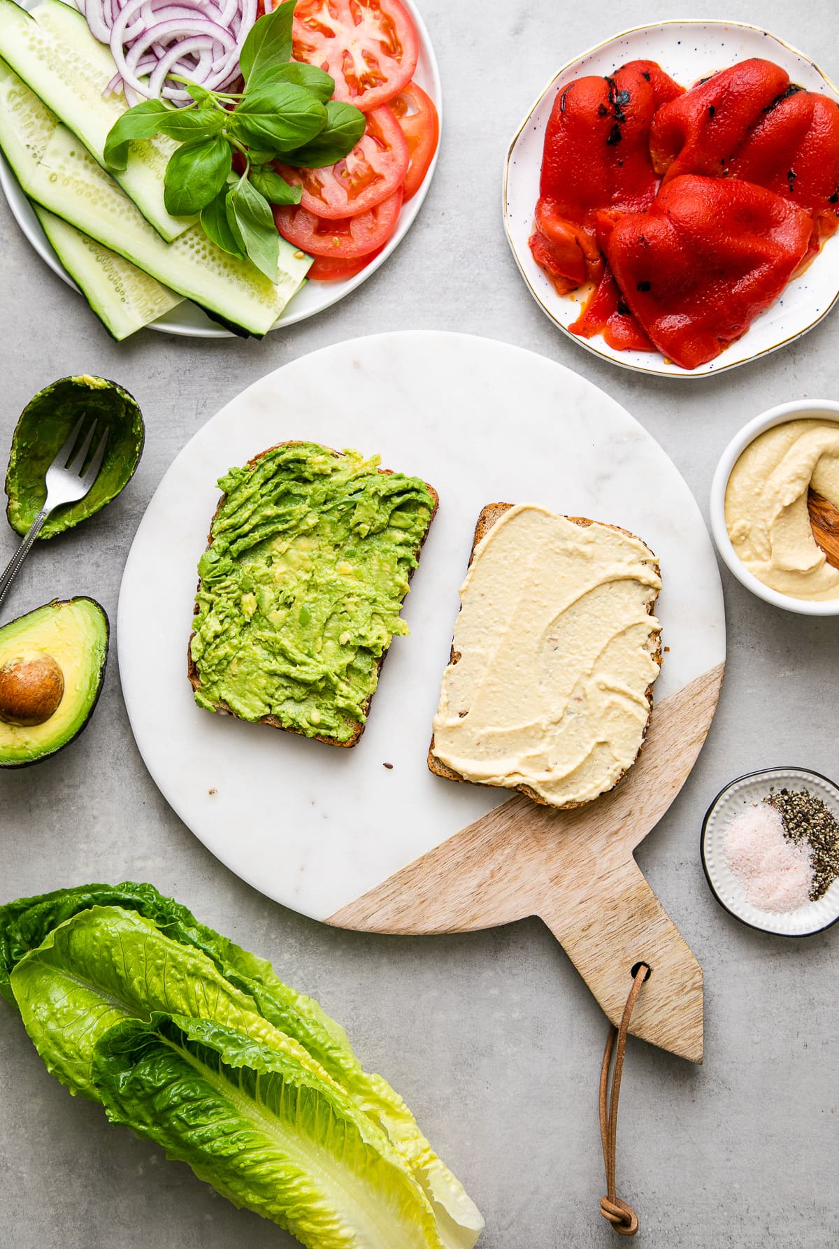 top down view showing the process of layer avocado and hummus on bread slices with items surrounding.